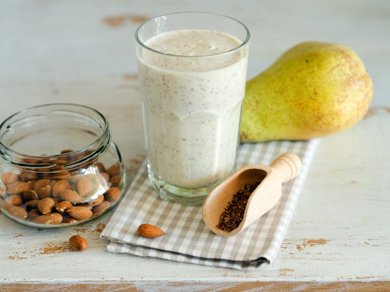 A glass with a creamy pear-almond smoothie. The glass is standing on a checkered kitchen towel. There is also a pear in the picture. Next to the glass is a glass bowl with almonds. Additionally, a small wooden shovel with a few linseeds can be seen.