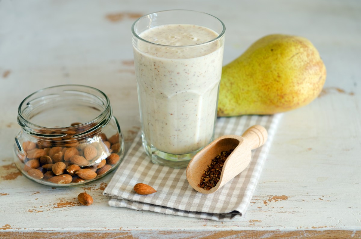 A glass with a creamy pear-almond smoothie. The glass is standing on a checkered kitchen towel. There is also a pear in the picture. Next to the glass is a glass bowl with almonds. Additionally, a small wooden shovel with a few linseeds can be seen.