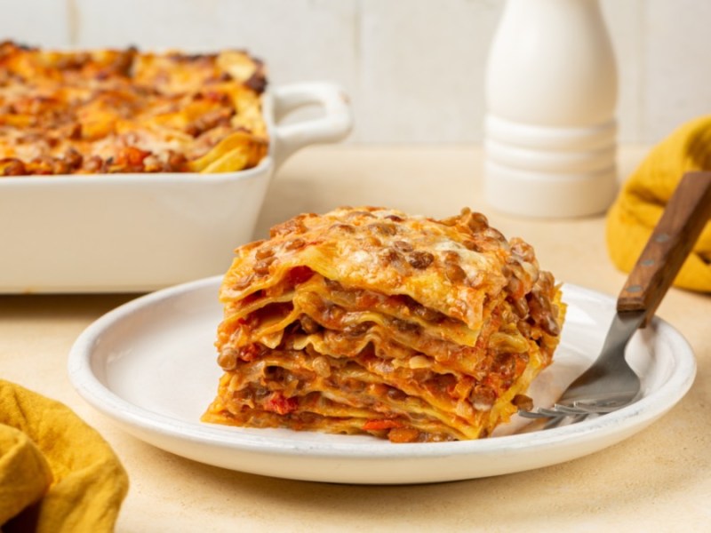 A slice of lentil lasagna on a plate. In the background, the rest can be seen in a baking dish.