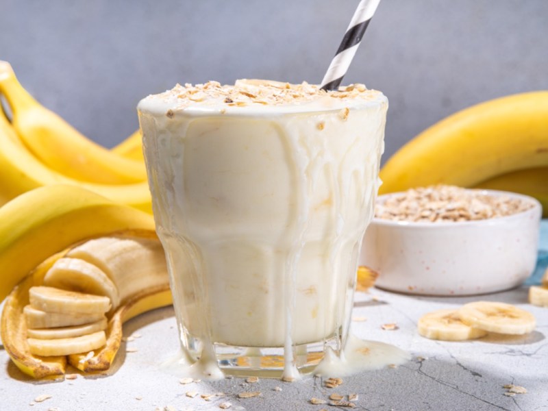 A glass of breakfast shake with bananas and oatmeal. In the background are bananas and a bowl of oatmeal.