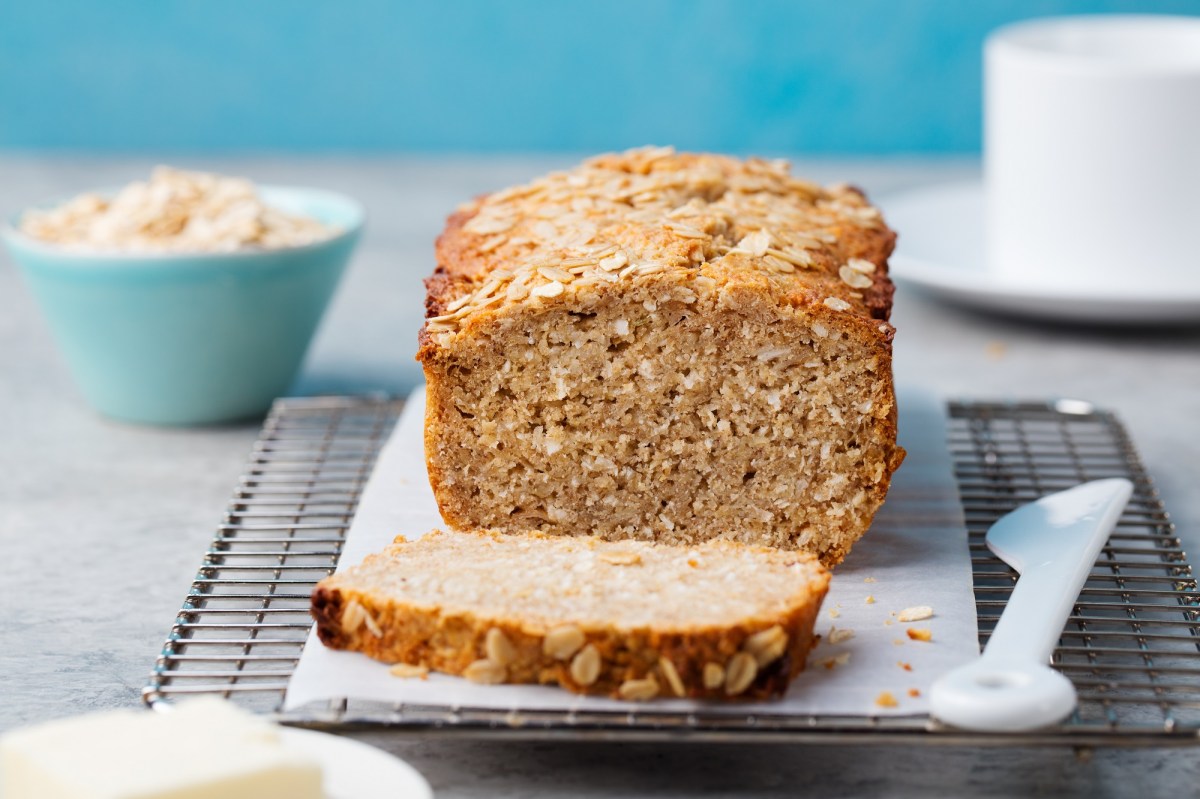A sliced oat bread on a board, next to it a white knife, in the background blurred a bowl with rolled oats.