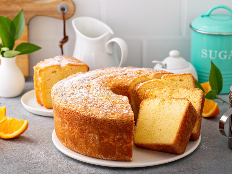 A partially sliced pound cake on a light gray countertop. Next to it are a sugar bowl, a pitcher, and a powdered sugar shaker.