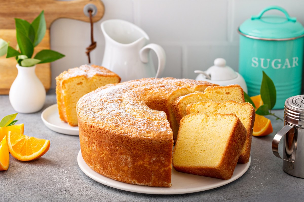 A partially sliced pound cake on a light gray countertop. Next to it are a sugar bowl, a pitcher, and a powdered sugar shaker.