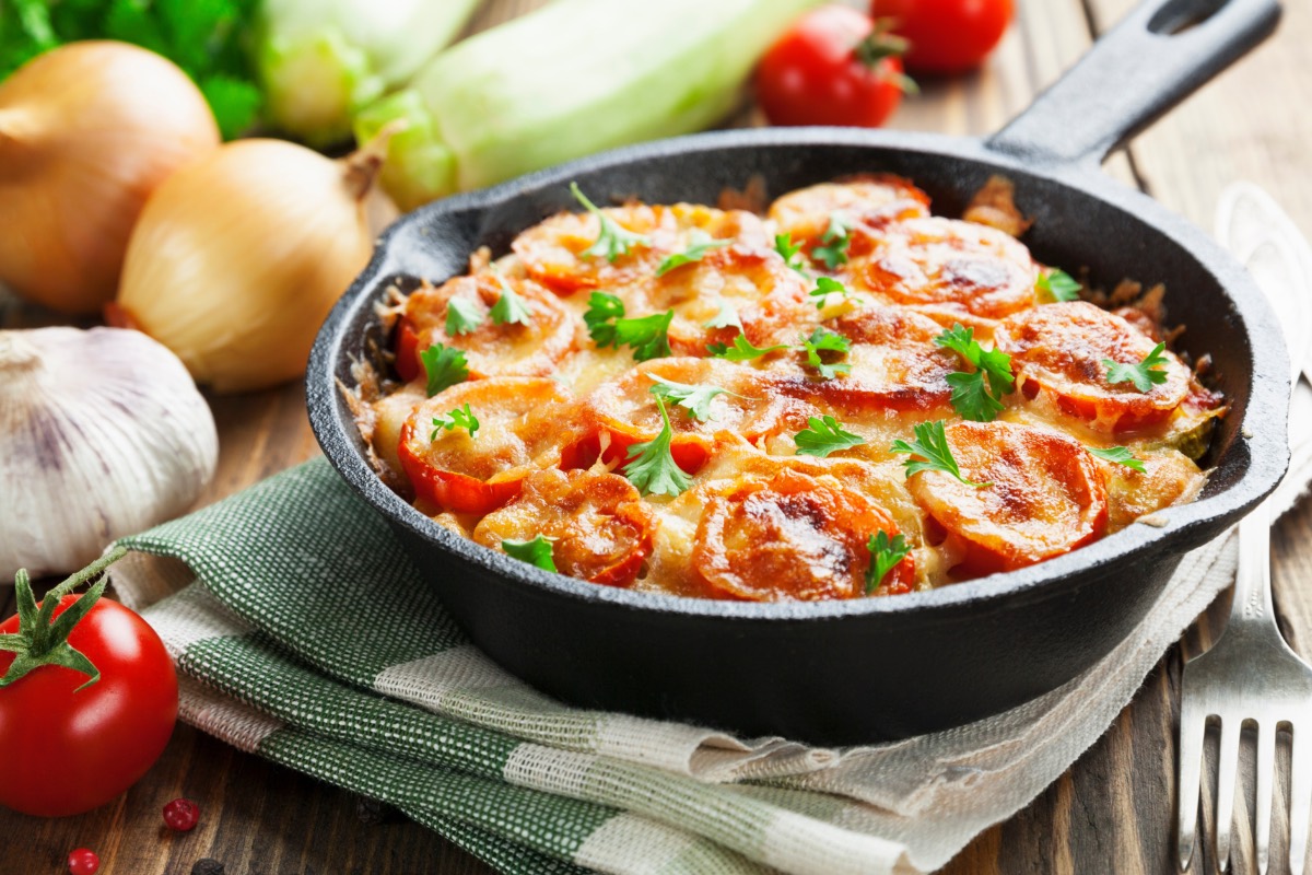 Tomato and zucchini casserole in an ovenproof pan on a dish towel. Fresh ingredients lie in the background.