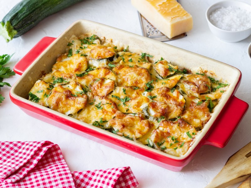 Zucchini casserole in a red casserole dish, garnished with chives. A piece of parmesan lies next to it.