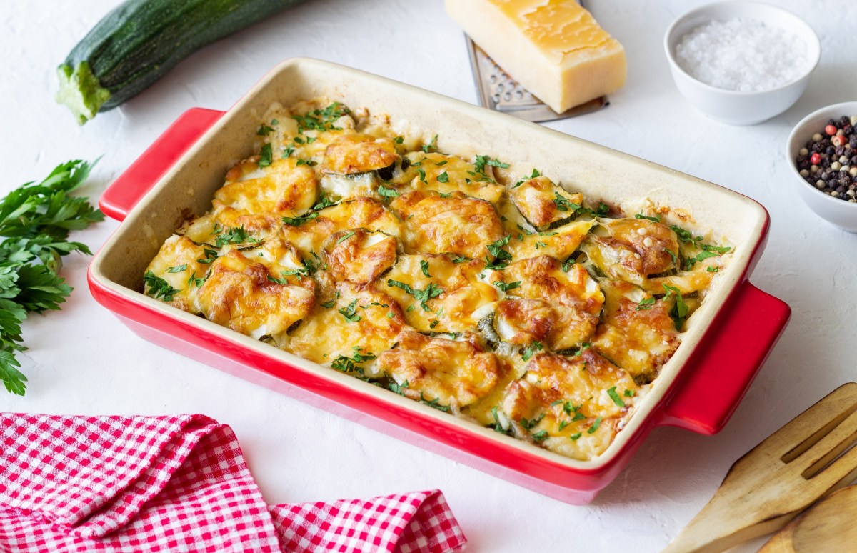 Zucchini casserole in a red casserole dish, garnished with chives. A piece of parmesan lies next to it.