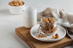 A clear glass sits on a table. Inside the glass, there is crunchy granola. The granola is golden brown and made of oats, nuts, and seeds. Some pieces are small, others are bigger. You can see the different textures.