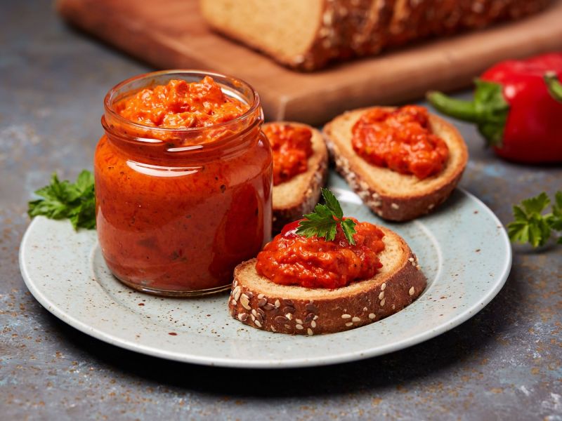 A glass of ajvar-feta dip stands on a plate, next to it are several slices of bread with the dip. In the background are bread and peppers as decoration.