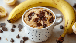 A banana mug cake sits on a light background. Chocolate pieces and a banana are scattered around the mug.