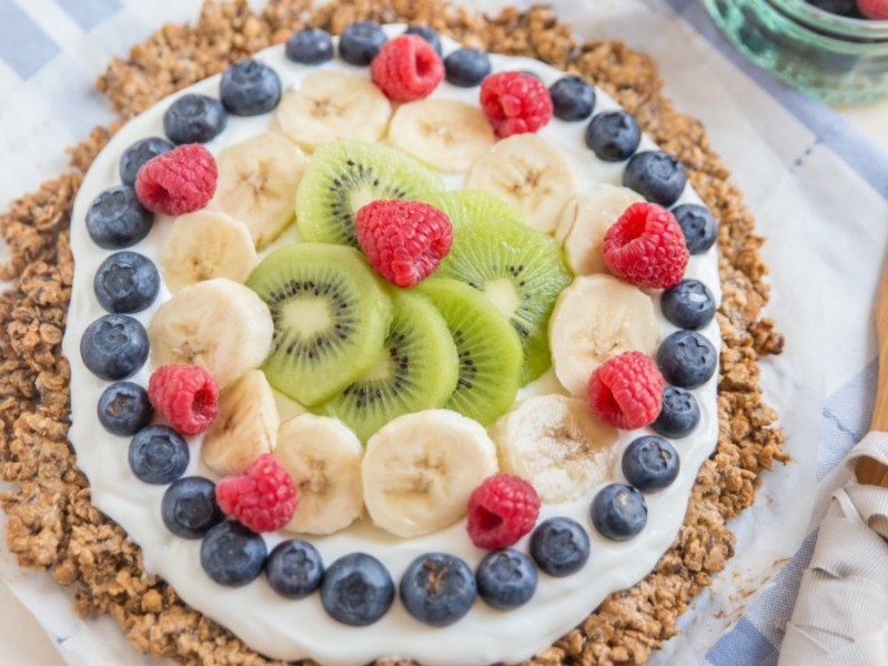 A breakfast pizza pictured from above with cream and a ring of bluebrries, with bananas and kiwi in the center.