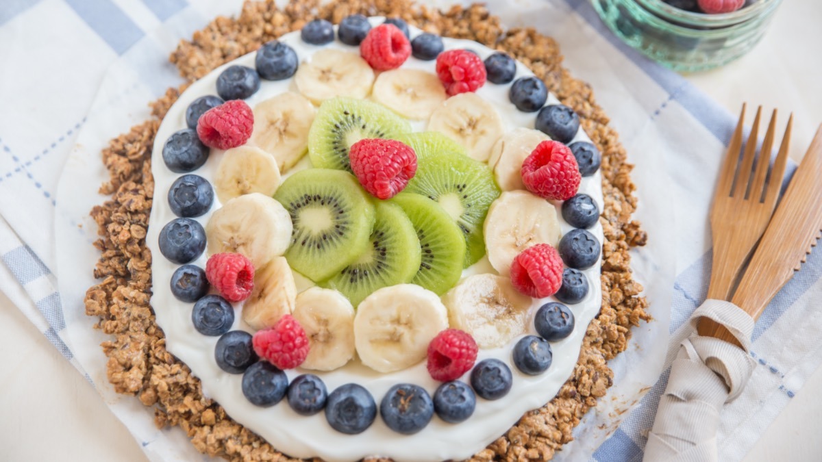A breakfast pizza pictured from above with cream and a ring of bluebrries, with bananas and kiwi in the center.