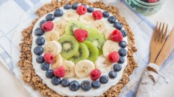 A breakfast pizza pictured from above with cream and a ring of bluebrries, with bananas and kiwi in the center.