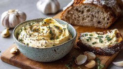 A bowl of caramelized garlic butter, which is on a wooden board with a sliced sourdough bread. Next to the bowl is a slice of bread spread with the butter. Around it are individual cloves of garlic as well as whole heads.