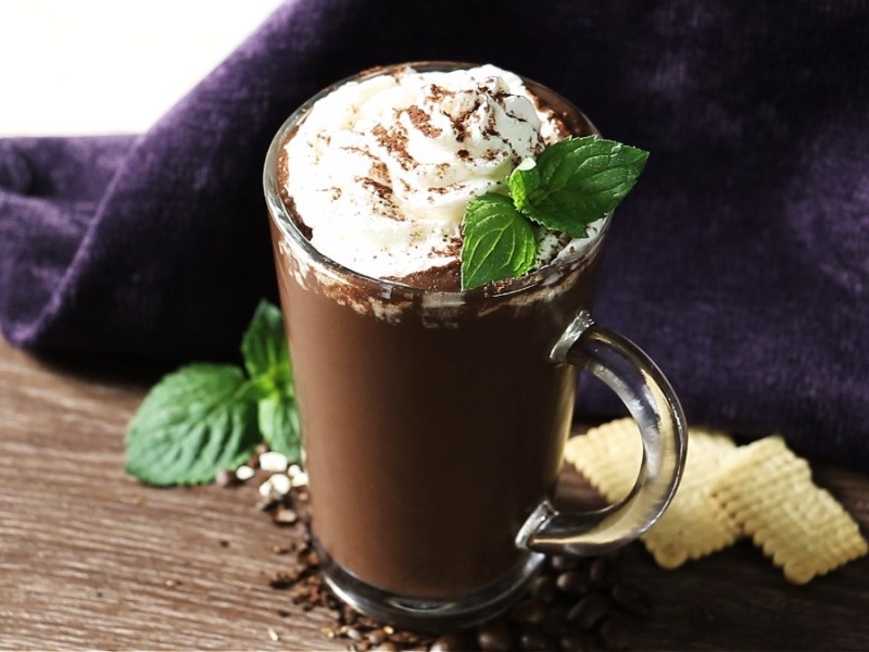 A glass of coffee smoothie sits on a wooden table. It’s topped with whipped cream and a mint leaf. The light brown smoothie contrasts with the white cream and green mint.