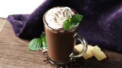A glass of coffee smoothie sits on a wooden table. It’s topped with whipped cream and a mint leaf. The light brown smoothie contrasts with the white cream and green mint.