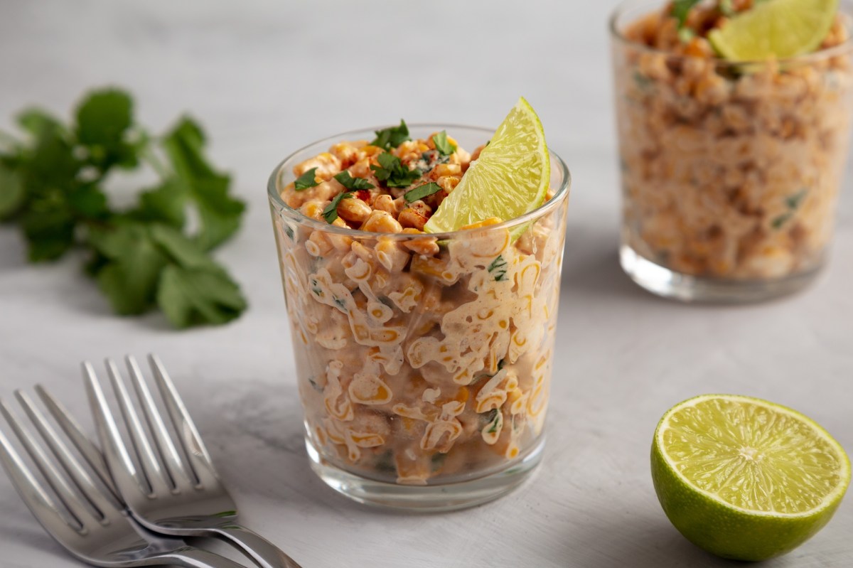 2 glasses of esquites, Mexican street corn salad, garnished with lime wedges and cilantro, next to two forks, fresh cilantro, and a halved lime.