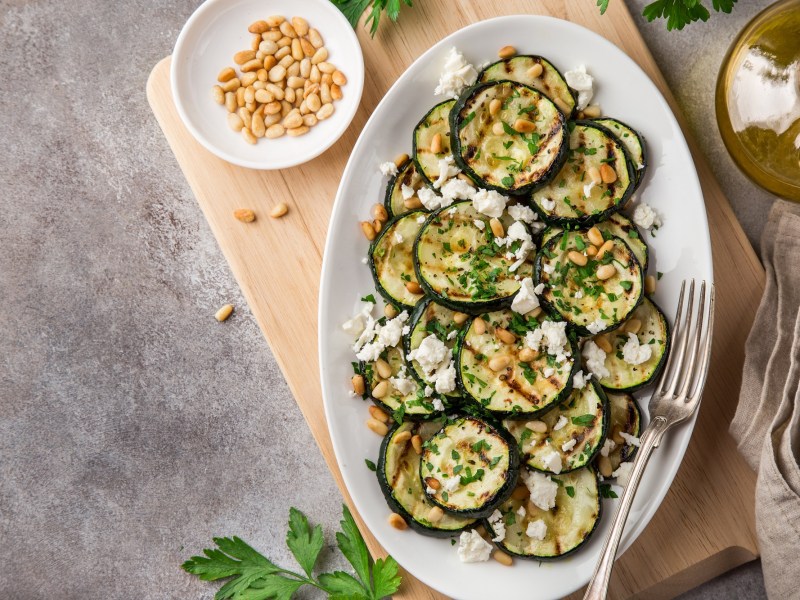 Sliced grilled zucchini sprinkled with feta and pine nuts on an oval plate, top view. Next to it chili flakes in a bowl, an oil bottle and other ingredients.
