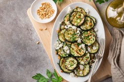 Sliced grilled zucchini sprinkled with feta and pine nuts on an oval plate, top view. Next to it chili flakes in a bowl, an oil bottle and other ingredients.