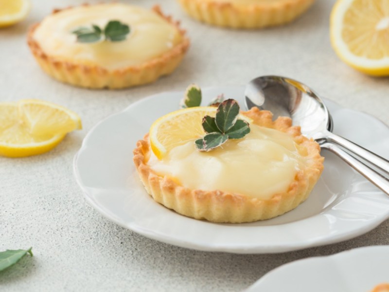 Two lemon curd tartlets on plates. Cutlery lies next to one, sliced lemons in the background.
