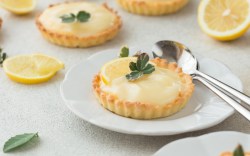 Two lemon curd tartlets on plates. Cutlery lies next to one, sliced lemons in the background.