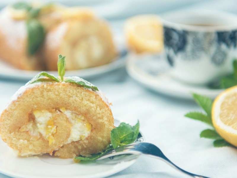 A slice of lemon swiss roll, decorated with mint, on a plate. A cup of coffee and the rest of the cake can be seen in the background.