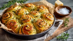 A round baking pan with savory herb rolls. The baking pan is on a wooden board decorated with a kitchen towel. Fresh herbs are scattered around. You can also see a piece of Parmesan and a small bowl of coarse sea salt.