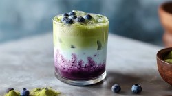 A glass of blueberry matcha latte. On the work surface you can still see a pile of matcha powder and a bowl of matcha powder. There are also a few individual blueberries lying around.