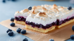 Several pieces of blueberry meringue sheet cake are arranged on a piece of baking paper. A few blueberries are scattered around as decoration.