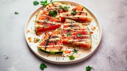 A plate with a few quarters of grilled watermelon, garnished with tahini, chili oil, black sesame seeds and fresh cilantro.