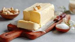 A wooden board with a block of vegan butter garnished with two cashews. In the background, there's a bowl of cashews. Next to the board is a small bowl of salt. A knife rests on the board.