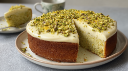 A plate with a round, sliced orange pistachio cake with ricotta frosting. In the background, you can see a plate with a piece of cake and a cup of coffee.