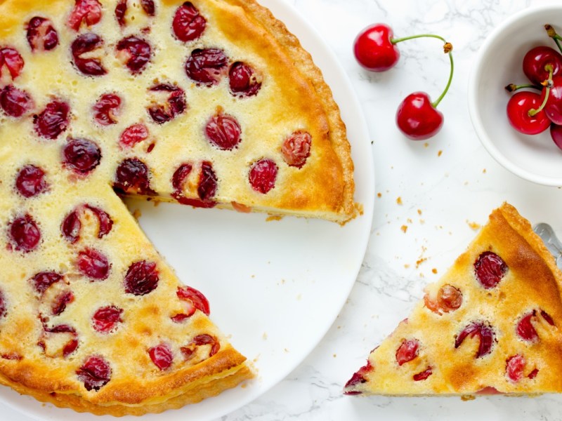 An overhead view of a Pudding-Cherry Cake with a slice cut out. The slice of cake and a bowl of fresh cherries are next to it.