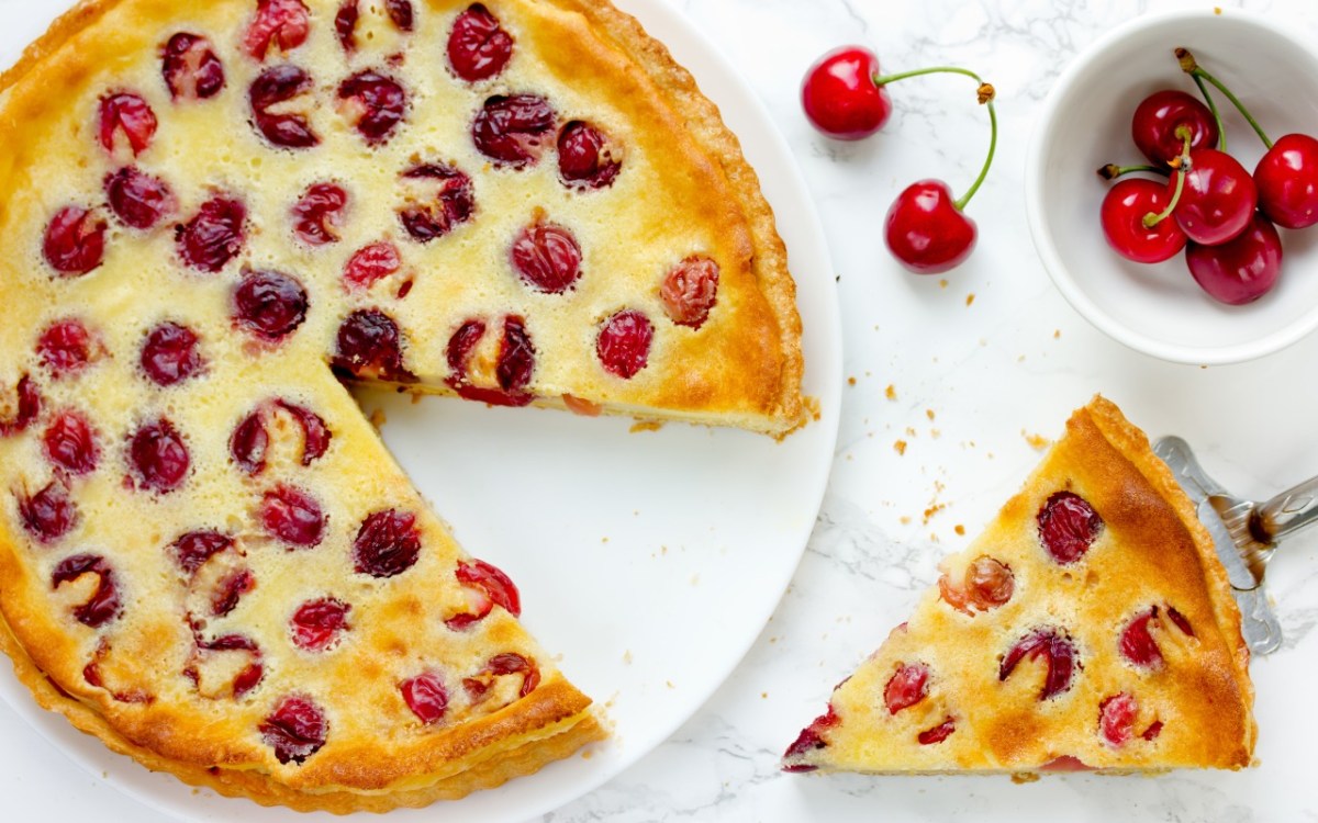 An overhead view of a Pudding-Cherry Cake with a slice cut out. The slice of cake and a bowl of fresh cherries are next to it.