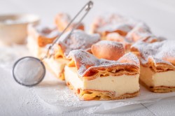 Puff pastry cake with vanilla custard on a white marble slab. On top is a sieve with which you can sprinkle the cake with powdered sugar.