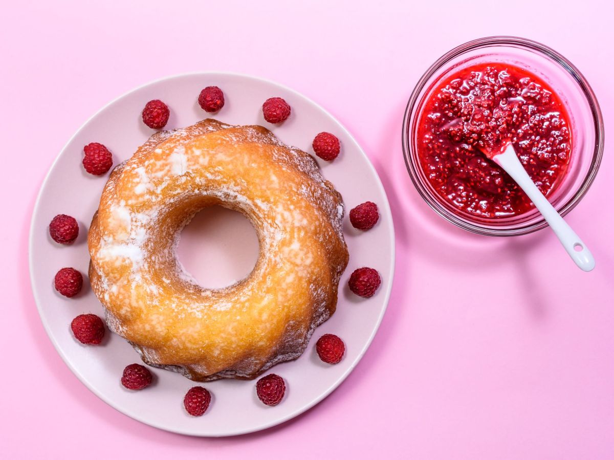 Raspberry lemon bundt cake, served on a plate and decorated with fresh raspberries. A small bowl of raspberry compote is next to it.