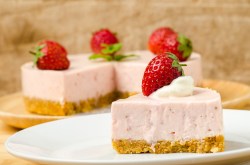 A piece of strawberry mousse cake, garnished with a strawberry, on a plate, with the cut cake in the background.