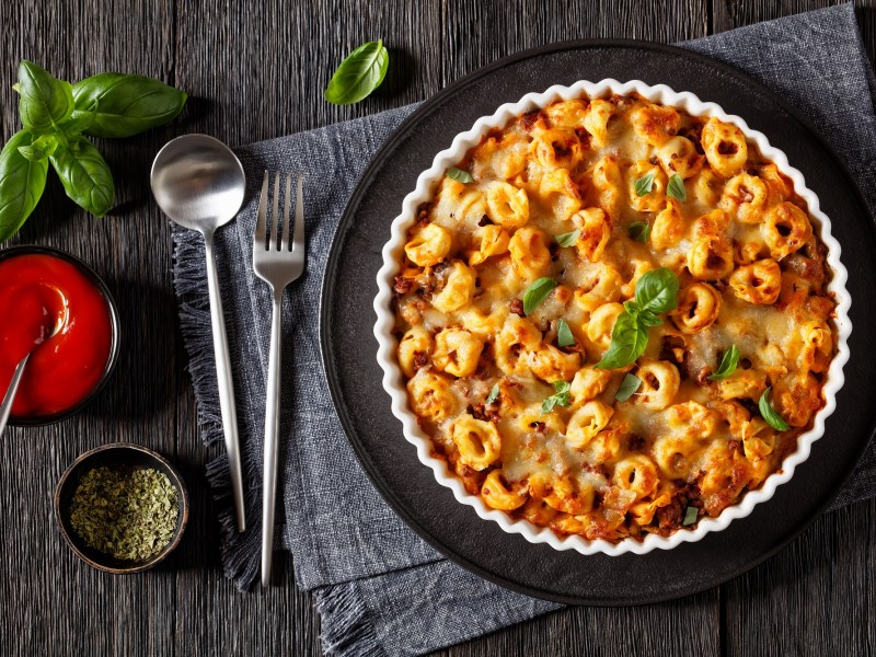 Tortellini casserole in a fluted baking dish on a black tray. Below is a gray placemat, spoon and fork, and tomato sauce. Overhead view.