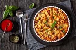 Tortellini casserole in a fluted baking dish on a black tray. Below is a gray placemat, spoon and fork, and tomato sauce. Overhead view.
