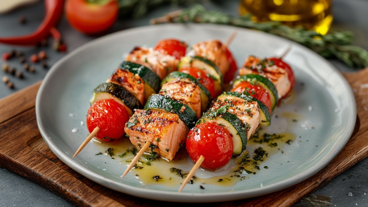 A plate with three zucchini salmon skewers marinated in a herb marinade. The plate is on a dark wooden board. In the background, you can see fresh ingredients like chili peppers, a tomato, sprigs of herbs, and a glass container with olive oil.