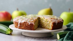 A round apple zucchini cake lying on a cake stand. Fresh apples and zucchini are scattered around.