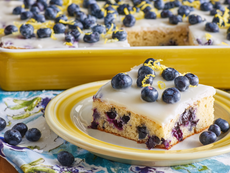 A piece of blueberry lemon sheet cake. The rest of the cake is in the background.
