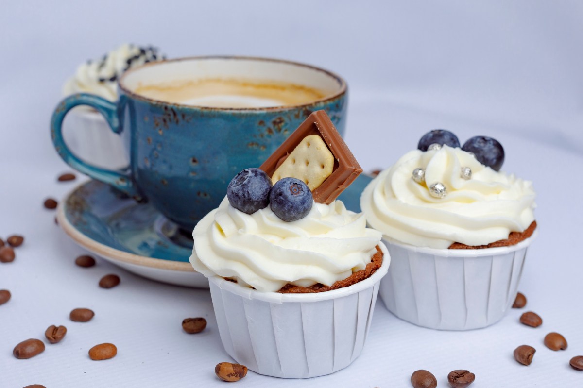 Two cappuccino cupcakes decorated with chocolate, blueberries, and sprinkles, with a blue cup of cappuccino in the background.