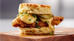 Chicken biscuit on a wooden block with dill pickle slices and sriracha mayo in a full frontal view with white lighting.