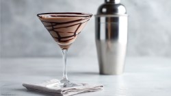 Chocolate martini on a cocktail napkin with cocktail shaker behind it with white lighting and a frontal view.
