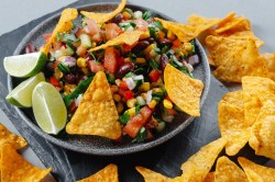 A bowl of Cowboy Caviar, a salad of beans, corn, onions, tomatoes and peppers, topped with lime wedges and tortilla chips. Close-up.