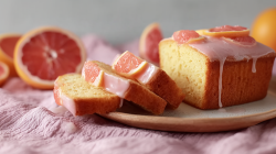 A sliced grapefruit yogurt loaf cake with a pink glaze and garnished with a few grapefruit slices. The cake is on a plate, which is on a pink cloth. In the background you can see whole and sliced grapefruits.