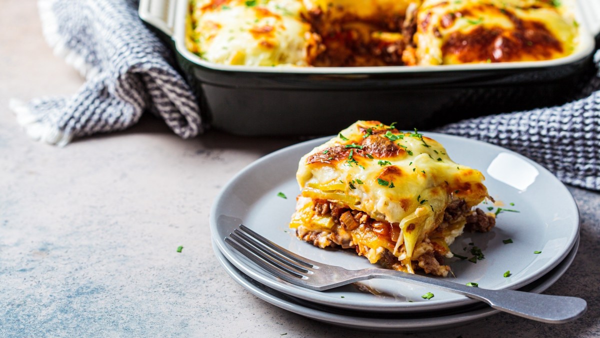 A piece of Greek Gyro Potato Casserole lies in front of a casserole dish with more casserole in it.