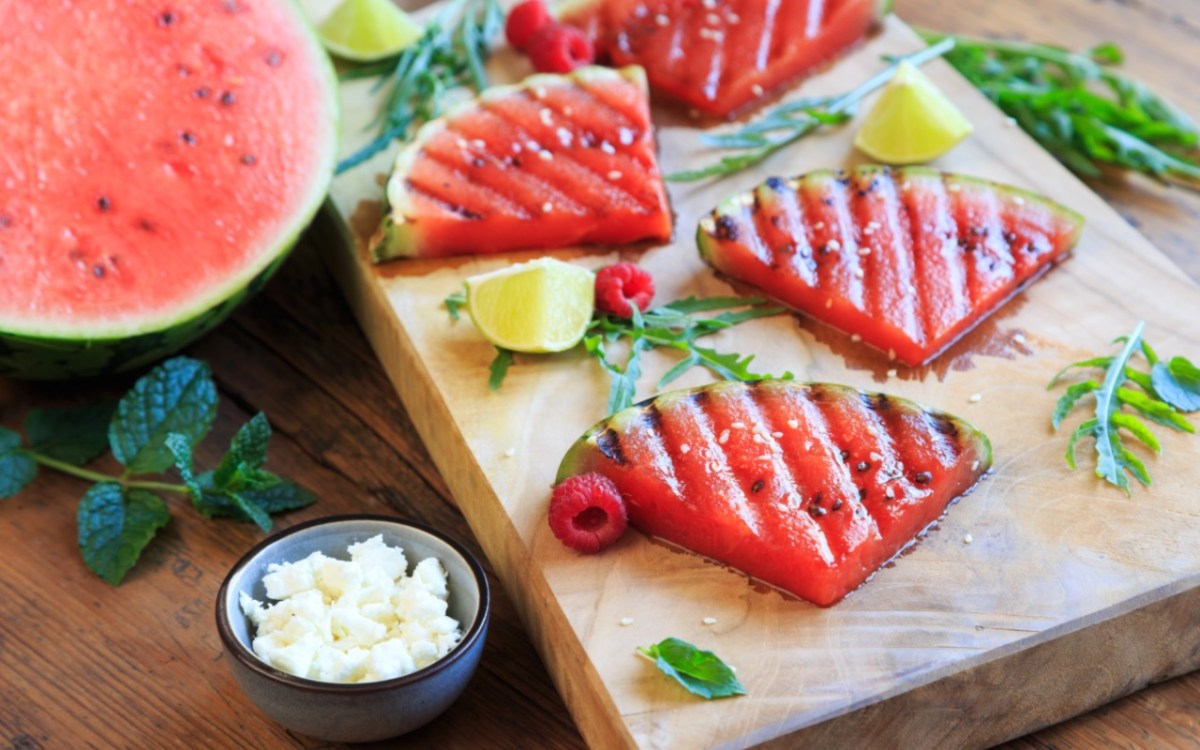 Grilled watermelon slices on a wooden board. Next to it is a bowl of feta.