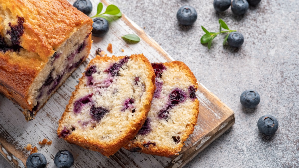 A lemon blueberry bread on a board, two slices cut off and lying next to it. Next to it are fresh blueberries and mint leaves.