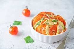 Mini vegetable gratin with tomato and zucchini in a white round baking dish, mini tomatoes next to it.